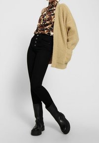 Béžový pletený cardigan navršený přes vzorovanou hnědo-černou halenku, doplněný o černé kalhoty s vysokým pasem a robustní černé kotníkové boty.