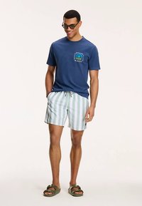 Marineblauw katoenen T-shirt met een logo; groen-wit gestreepte zwemshorts; bruine sandalen; zonnebril; comfortabel model, casual ontwerp.
