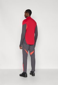 Conjunto deportivo que incluye una camiseta de manga larga roja y gris y pantalones grises con detalles naranjas, confeccionados con una tela suave y ligera.