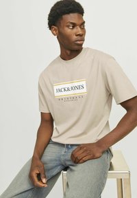 Beige Baumwoll-T-Shirt mit einem zentralen Logo und dem Text "JACK & JONES", kurzen Ärmeln und einer lockeren Passform, kombiniert mit blauen Jeans.