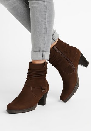 Brune ankelstøvler med sidelås og knapdetalje, båret med lysegrå optrukne skinny jeans på hvid baggrund.