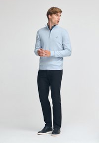 Ljusblå tröja med dragkedja, ribbade ärmslut och en liten logotyp, tillsammans med svarta byxor och mörka sneakers. Slät textur.