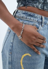 Hõbedane ketigaelne käevõru, millel on dekoratiivne ripats, kantud käel, mis resting kergetes sinistes kulunud teksades kollaste aktsentidega.