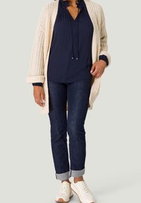 Cardigan en maille crème porté sur un chemisier bleu marine à col volanté, associé à un jean en denim foncé et des baskets blanches avec des accents beige.