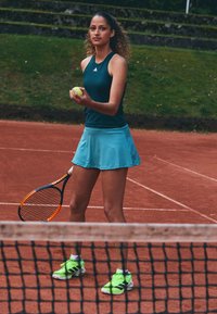 Giocatore di tennis in una canotta turchese e una gonna turchese, con in mano una pallina da tennis gialla. Scarpe verde neon con dettagli neri. Racchetta da tennis in mano.