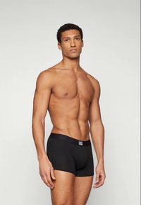 Muscular man standing, wearing black boxer briefs with a logo waistband, against a plain light background.
