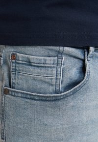 Svetle modré džíny z denimu s zadní kapsou a malým měděným nýt. Textura je mírně opotřebovaná s viditelnými švy.