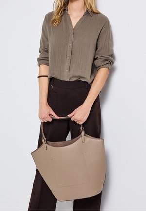 Vrouw draagt een bruin overhemd met knopen en donkere broek, houdt een grote beige leren handtas met twee hengsels voor een witte achtergrond.