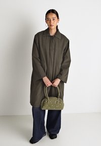Oversized olive green coat with a collar, paired with dark blue wide-leg jeans. A small green handbag with embellishments in hand.