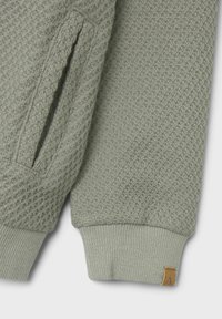 Svetlozelený sveter s textúrovaným vzorom, rebrovanými manžetami a bočnou vreckom. Obsahuje béžovú etiketu s písmenom "A" na leme.