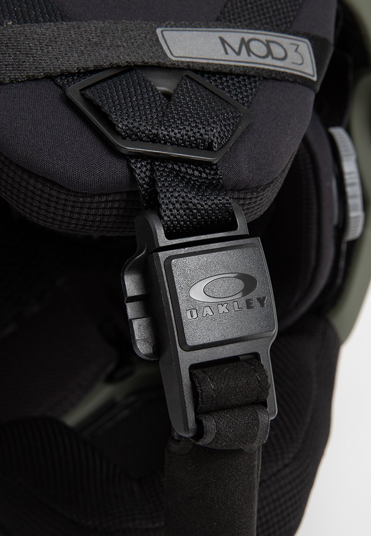 Czarny pasek hełmu narciarskiego z okrągłą czarną klamrą z logo Oakley; teksturowany materiał i regulowane taśmy zapewniające bezpieczne dopasowanie.