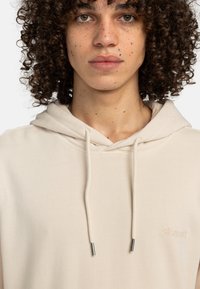 Sudadera beige con cordón y terminales plateados, que presenta un pequeño logo en relieve. Tejido suave y diseño informal.