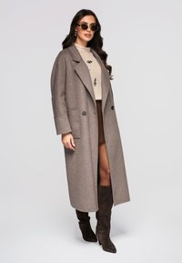 Long manteau marron en mélange de laine avec une coupe décontractée, deux boutons et des poches latérales. Porté avec des bottes hautes foncées jusqu'au genou et un haut clair.