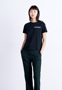 Zwarte katoenen T-shirt met korte mouwen, met een grafische print op de borst. Draagt een donkere groene broek en wordt weergegeven tegen een witte achtergrond.