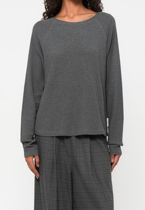 Frau trägt dunkelgraues langärmeliges Hemd und grau kariere weit geschnittene Hose, steht vor einem schlichten weißen Hintergrund.