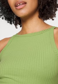 Lähikuva kiharoista hiuksista naisesta, joka käyttää ribbikuvioista vihreää hihattomaa toppia, esittäen kaulaa, hartioita ja huulia.