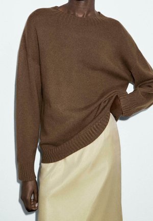 Personne portant un pull en maille brun à manches longues et une jupe beige, une main rangée dans la poche de la jupe, sur un fond uni.