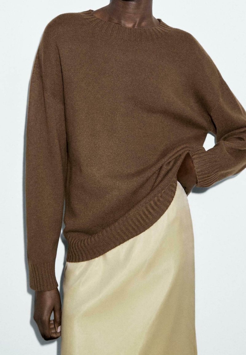 Personne portant un pull en maille brun à manches longues et une jupe beige, une main rangée dans la poche de la jupe, sur un fond uni.