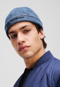 Denim beanie with a stitched brim and embossed logo in black. The wearer is in a blue bomber jacket, with visible stitching details.