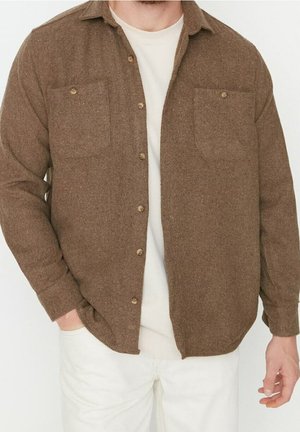 Homme portant une chemise marron à boutons par-dessus un t-shirt blanc avec un pantalon blanc, une main dans la poche, sur un fond blanc uni.