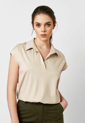 Beige Bluse mit Kragen und kurzen Ärmeln, glattem Stoff und einer teilweisen Knopfleiste. Kombiniert mit grünen Hosen. Neutraler Hintergrund.