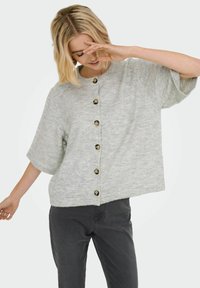 Femme portant un cardigan boutonné à manches courtes gris clair et un pantalon gris foncé, souriant avec une main près de son visage, sur fond blanc.