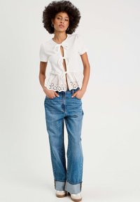 Top blanco de manga corta con un dobladillo peplum, que presenta tres lazos frontales y detalles de ojales; combinado con jeans de tiro alto de color azul claro.