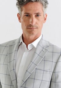 Jasnoszary blezer o subtelnym wzorze w kratę, wykonany z teksturowanej tkaniny. Noszony na białej koszuli z kołnierzykiem typu spread i zapięciem na guziki.