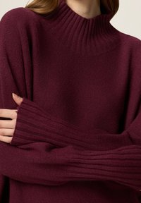 Pull col montant bordeaux en tissu doux, avec des poignets côtelés et une coupe décontractée, surface texturée et manches longues.