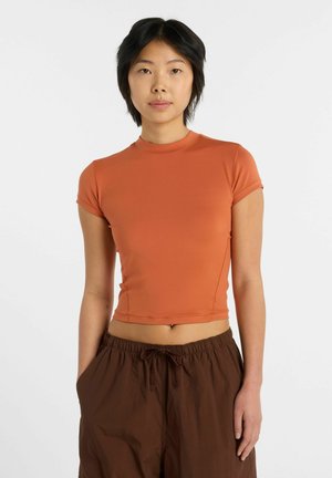 Person med kort sort hår iført en orange tætsiddende kortærmet top og løse brune bukser, stående mod en ensfarvet lys baggrund.