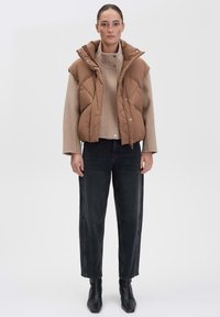 Braune Steppweste mit hohem Kragen, über einem beigen Pullover getragen, kombiniert mit schwarzen geraden Jeans und Stiefeln bis zum Knöchel. Glatte Textur, gestepptes Design.