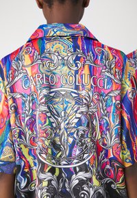 Camicia a maniche corte colorata con un vivace motivo astratto, dettagli floreali e grafici bianchi intricati, e il testo "CARLO COLUCCI" sul retro.