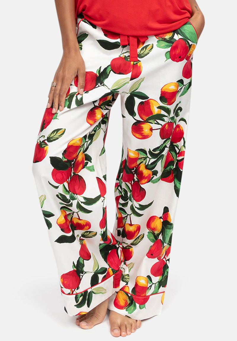 Pantalon blanc ample imprimé de fruits rouges et jaunes et de feuilles vertes, porté par une personne en haut rouge, debout pieds nus, mains dans les poches.