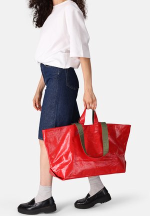 Red, glossy tote bag with green handles, large rectangular shape, smooth texture, and minimalist design, held by a person in denim skirt.