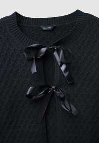 Cardigan nero in maglia testurizzata con colletto a coste e due fiocchi in raso nero legati all'apertura frontale, etichetta di dimensione small visibile.