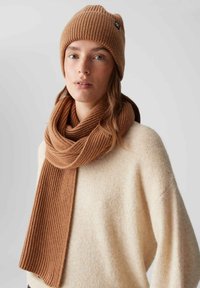 Donna che indossa un maglione beige, un berretto di maglia a coste marrone e una sciarpa coordinata su uno sfondo chiaro uniforme.