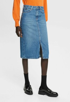 Personne portant une jupe en denim bleu mi-longue avec une fente à l'avant, des chaussettes noires, des mocassins noirs brillants et un haut orange à manches longues.