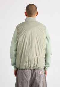 Hellgrüne Steppweste mit hohem Kragen und elastischer Säume, getragen über einem hellblau-grünen Sweatshirt, kombiniert mit grauen cordhosen.