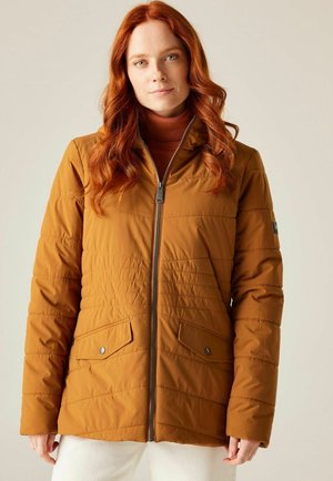 Kobieta z czerwonymi włosami w złocistej pikowanej kurtce na zamek i białych spodniach, stojąca na tle neutralnego tła.