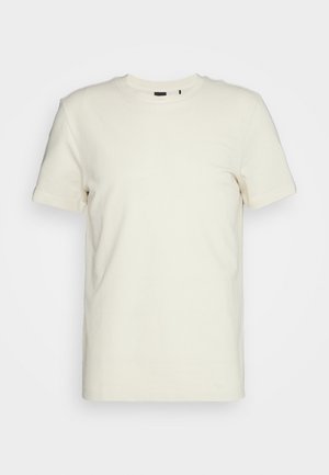 T-shirt a maniche corte di colore crema, colletto rotondo, tessuto testurizzato con un motivo sottile, con un piccolo etichetta nera vicino al collo.