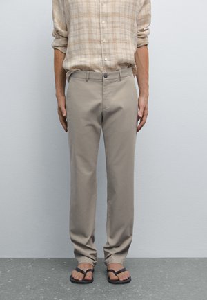 Beige cotton pants with a straight fit, featuring a front button closure. Worn with black flip-flops, and paired with a beige plaid shirt.
