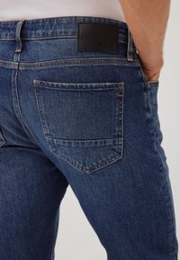 Jeans de mezclilla azul oscuro con un ajuste clásico, que cuentan con una parche de cuero negro en la parte trasera de la cintura y un bolsillo trasero con detalle de costura.