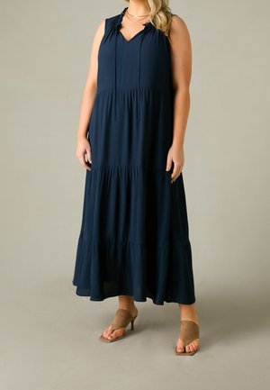 Maxikjole i marineblå med V-udskæring, detaljer med rynkede skuldre og lagdelt flæsekant. Stylet med lysebrune hælede mule-sko.