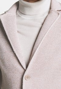 Světle růžový, žebrovaný svrchní oděv s klopovým límcem a zapínáním na jeden knoflík, vrstvený přes béžový rolák. Jemná textura, minimalistický design.