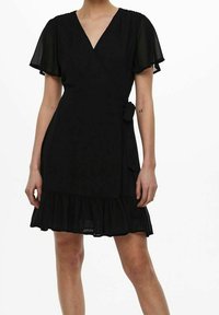 Donna che indossa un vestito nero avvolgente con maniche a sbuffo e orlo arricciato, legato in vita, su uno sfondo bianco semplice.