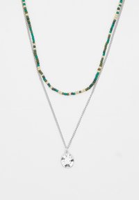 Geheel van kralen halsketting met groene, beige en blauwe kralen, gelaagd boven een zilveren ketting met een hanger met een gravure ontwerp.