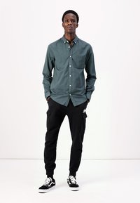 Camicia button-up verde scuro con tasca sul petto, abbinata a pantaloni cargo neri e sneakers nere con accenti bianchi. Design semplice, texture liscia.