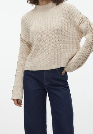 Vrouw draagt een beige gebreide trui met opengewerkt detail op de mouwen en donkeblauwe, hooggetailleerde jeans, hand in de zak.