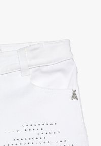 Gros plan sur une poche de pantalon en jean blanc avec passant de ceinture, détail métallique argenté en forme de mouche et petits clous argentés éparpillés sous la poche.