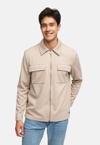 Beige lynlåsjakke med krave, struktureret stof, to brystlommer og lange ærmer, parret med lyseblå jeans.